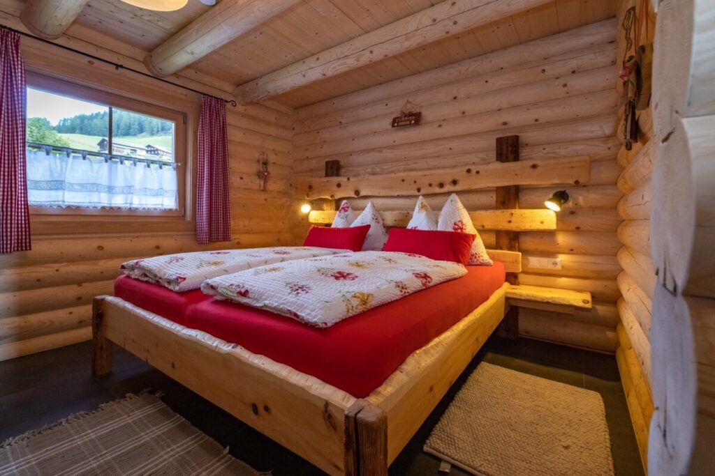 Schlafzimmer der Alm mit Holzwänden, Doppelbett und Leselampen, sowie Ausblick auf die umgebende Landschaft.