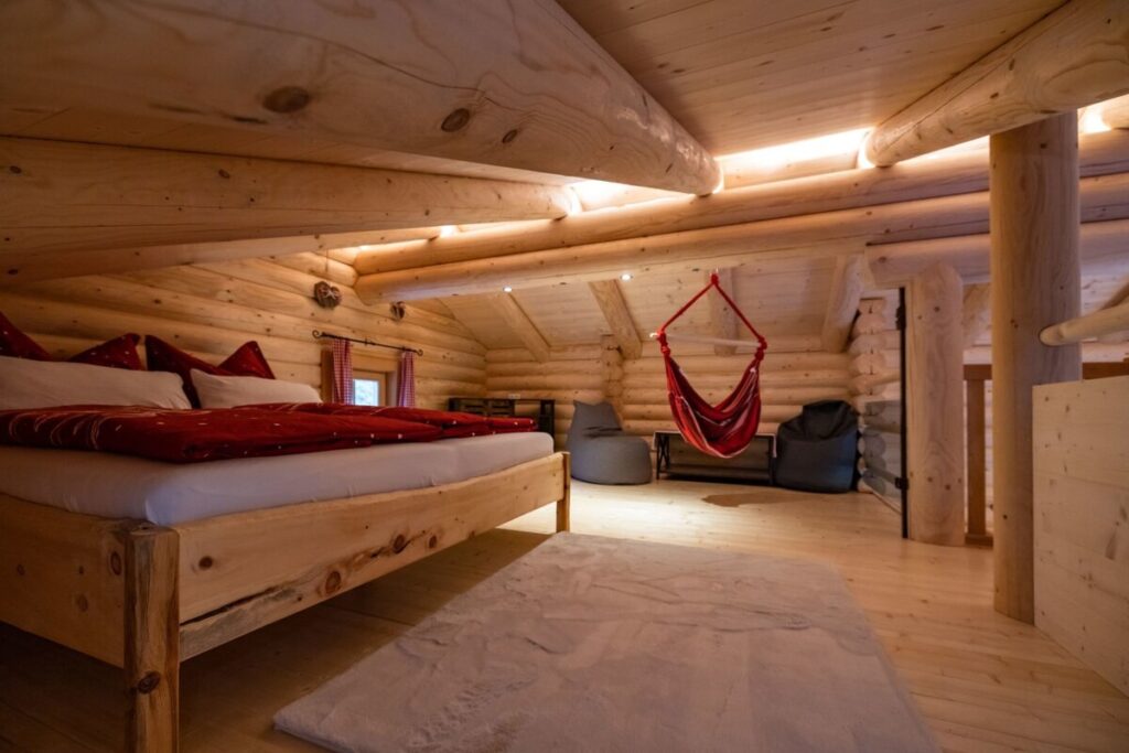 Schlafbereich in der Alm mit Holzbett, Hängesessel und massiven Holzbalken.